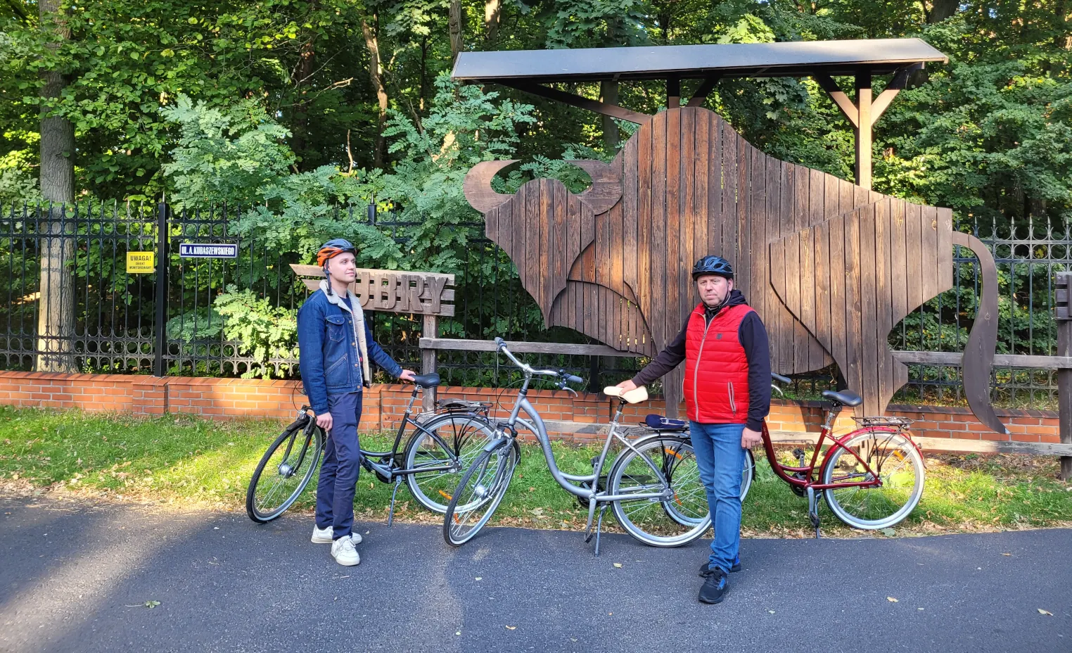 Dwoje rowerzystów w kaskach stojących z rowerami miejskimi przed drewnianą rzeżbą w kształcie żubra na tle parku w Gołuchowie. Zapraszamy do agroturystyki "Pod Bocianem"